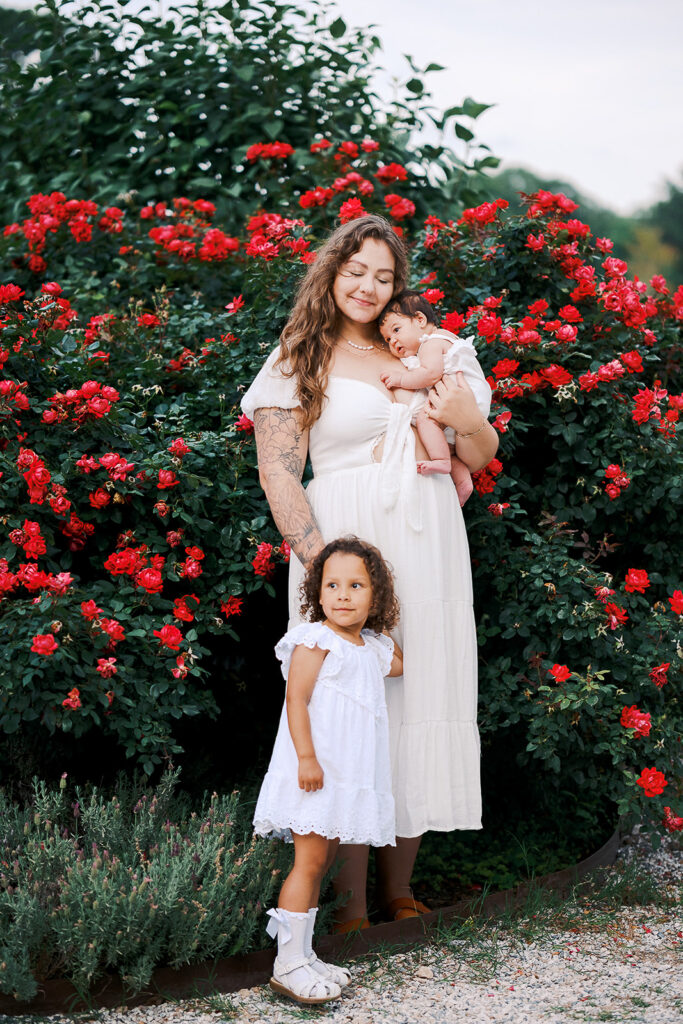 mom holding infant with toddler in front of her with flowers in the background