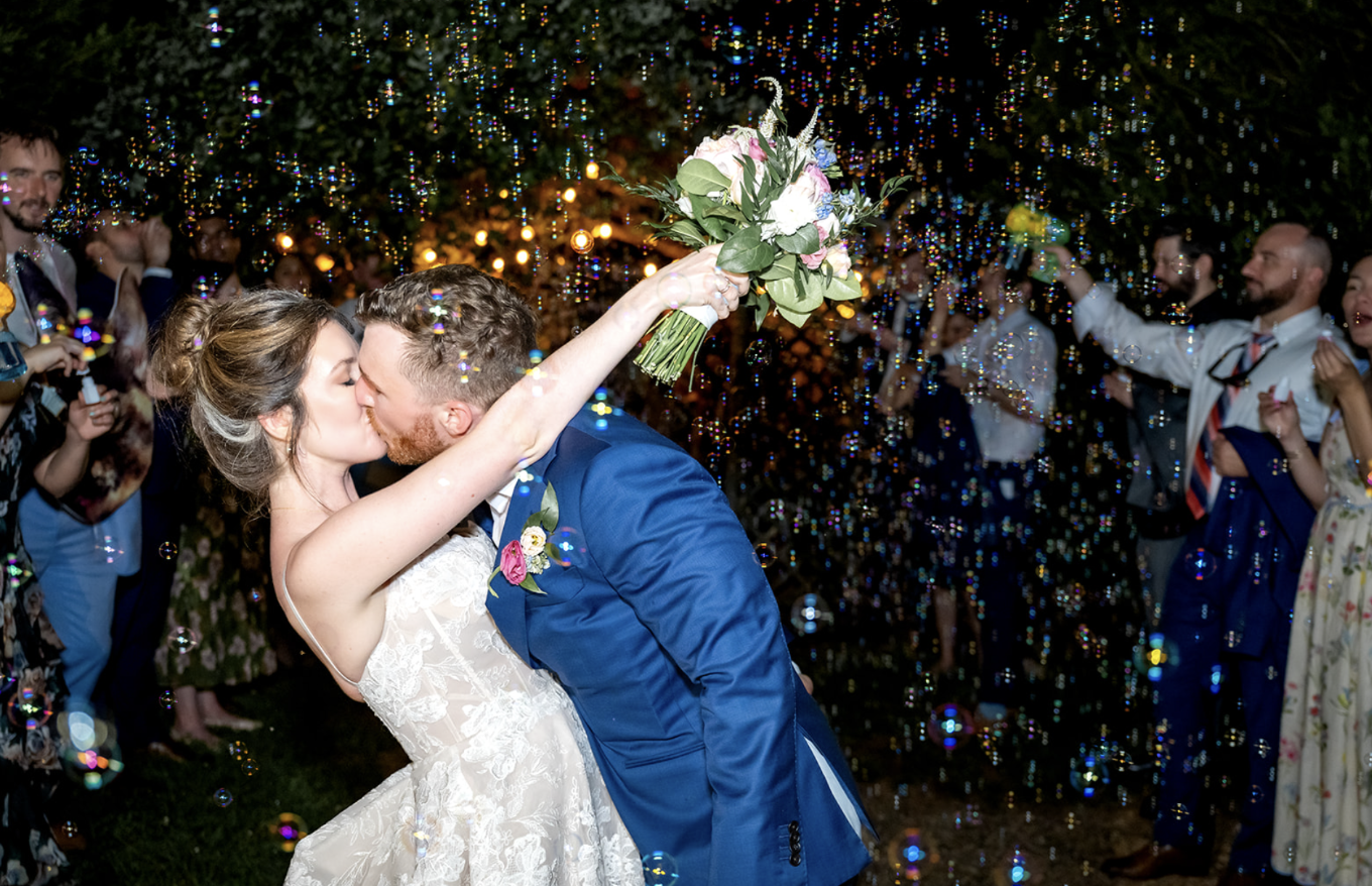 a couple, one member in a dress holding up a bouquet, and one in a suit, kiss in the night surrounded by bubbles.