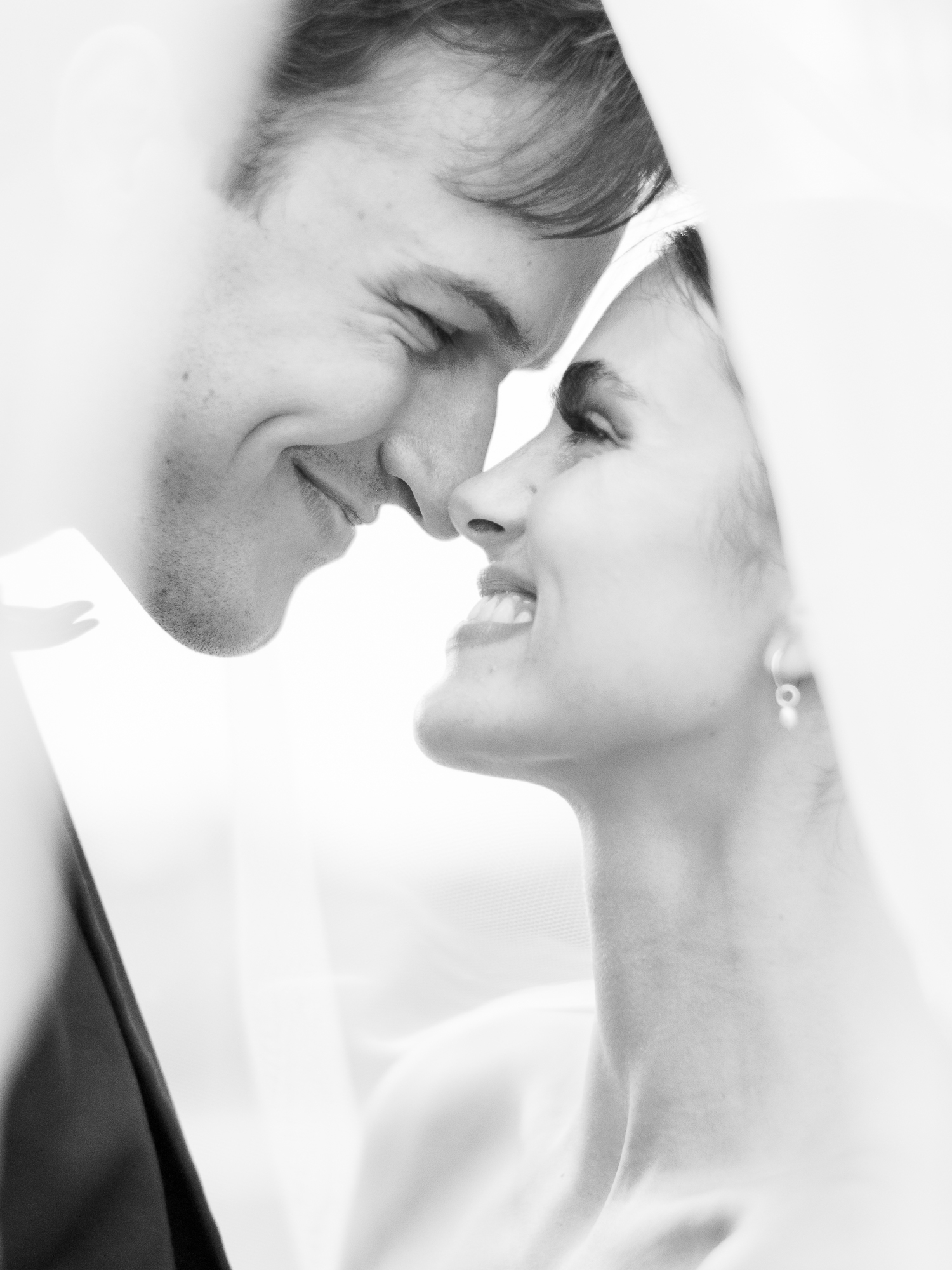 Close-up black and white portrait of bride and groom smiling under the veil, capturing an emotional luxury wedding moment.
