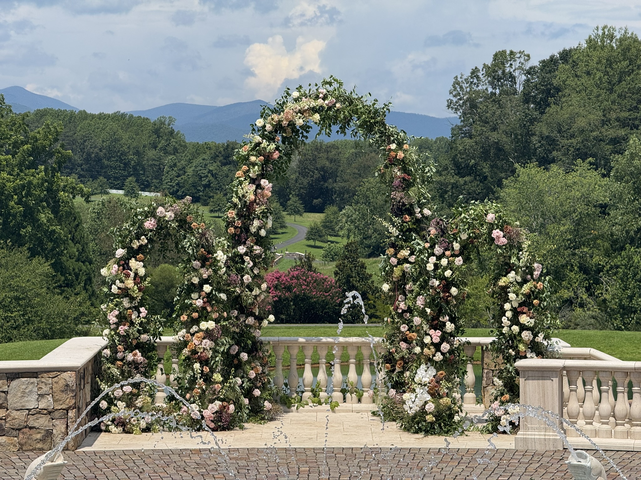 Large garden-style wedding ceremony arch covered in lush greenery and pastel floral arrangements overlooking mountains, manicured grounds, and an outdoor fountain.