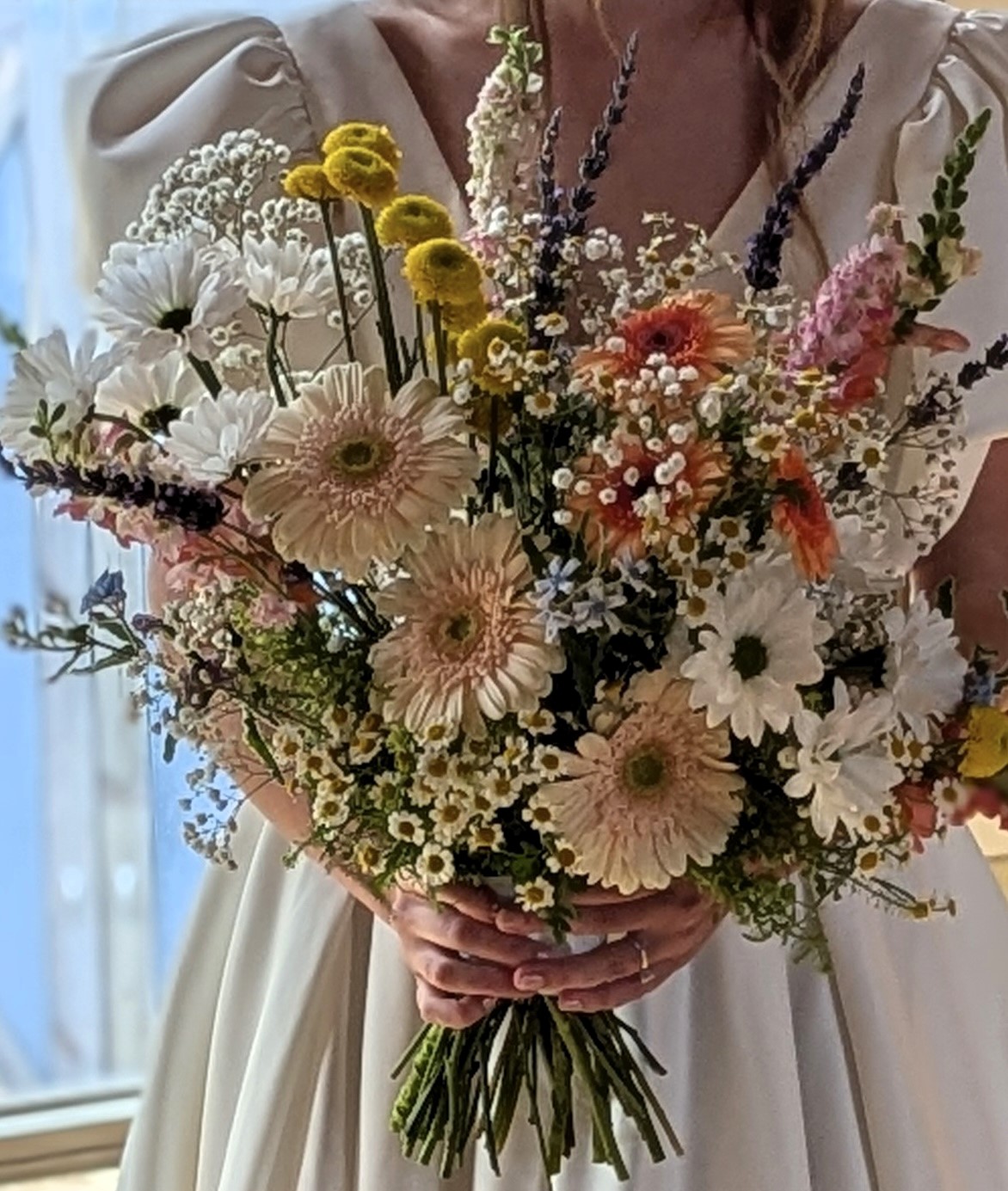 Bride holding a whimsical wildflower-style bouquet featuring blush gerbera daisies, white daisies, chamomile flowers, billy balls, snapdragons, and pastel seasonal blooms.